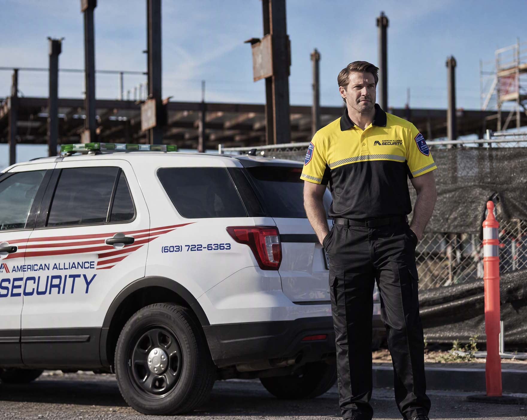 An American Alliance Security officer standing beside a marked patrol vehicle at a steel-frame industrial site