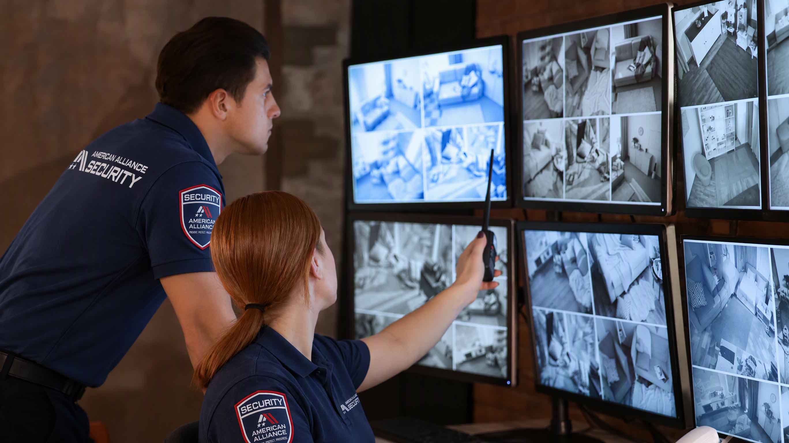 Two American Alliance Security operators watching a wall of camera monitors, one pointing at a feed with a radio