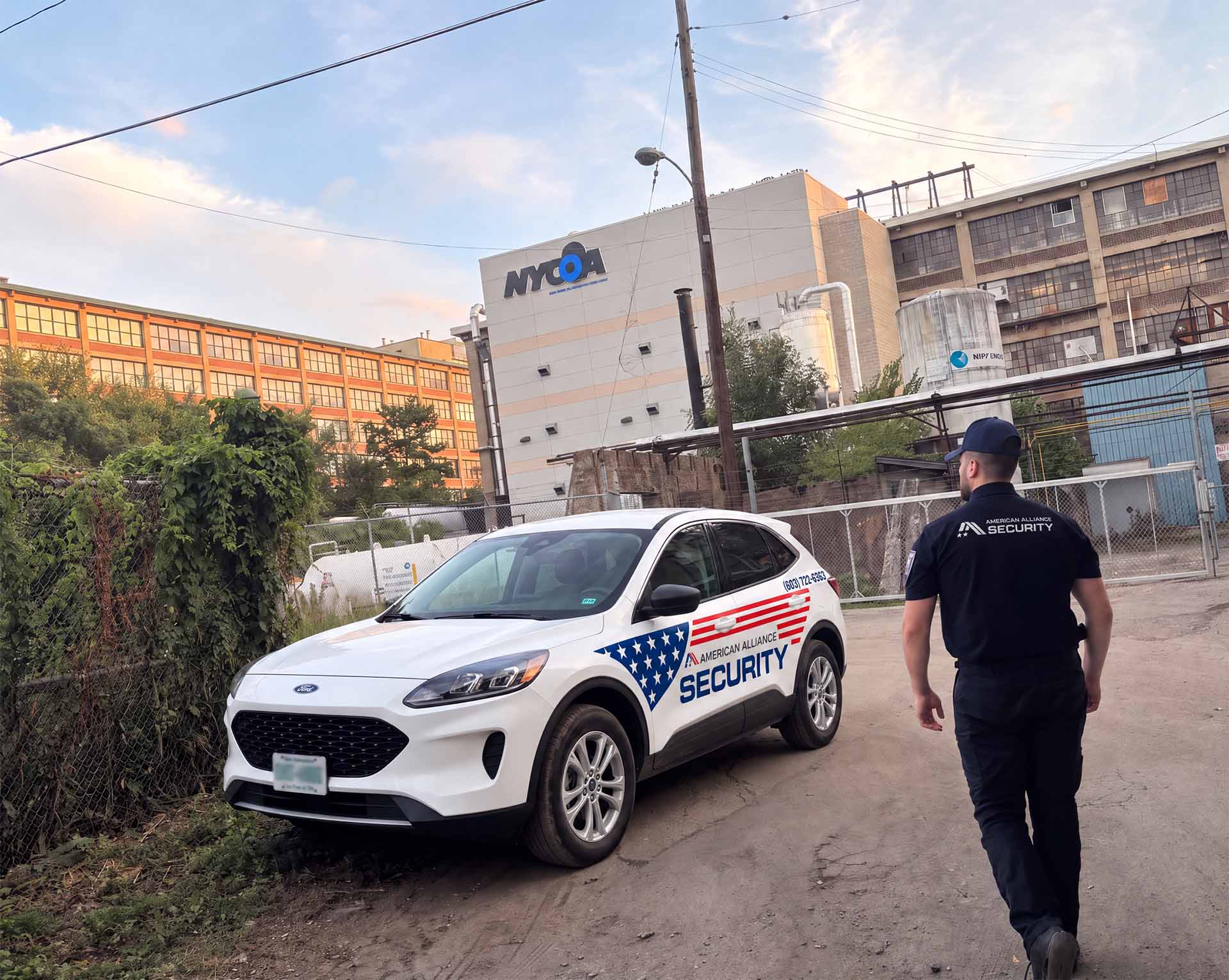 An American Alliance Security officer walking past a marked patrol vehicle along the fenced perimeter of an industrial plant