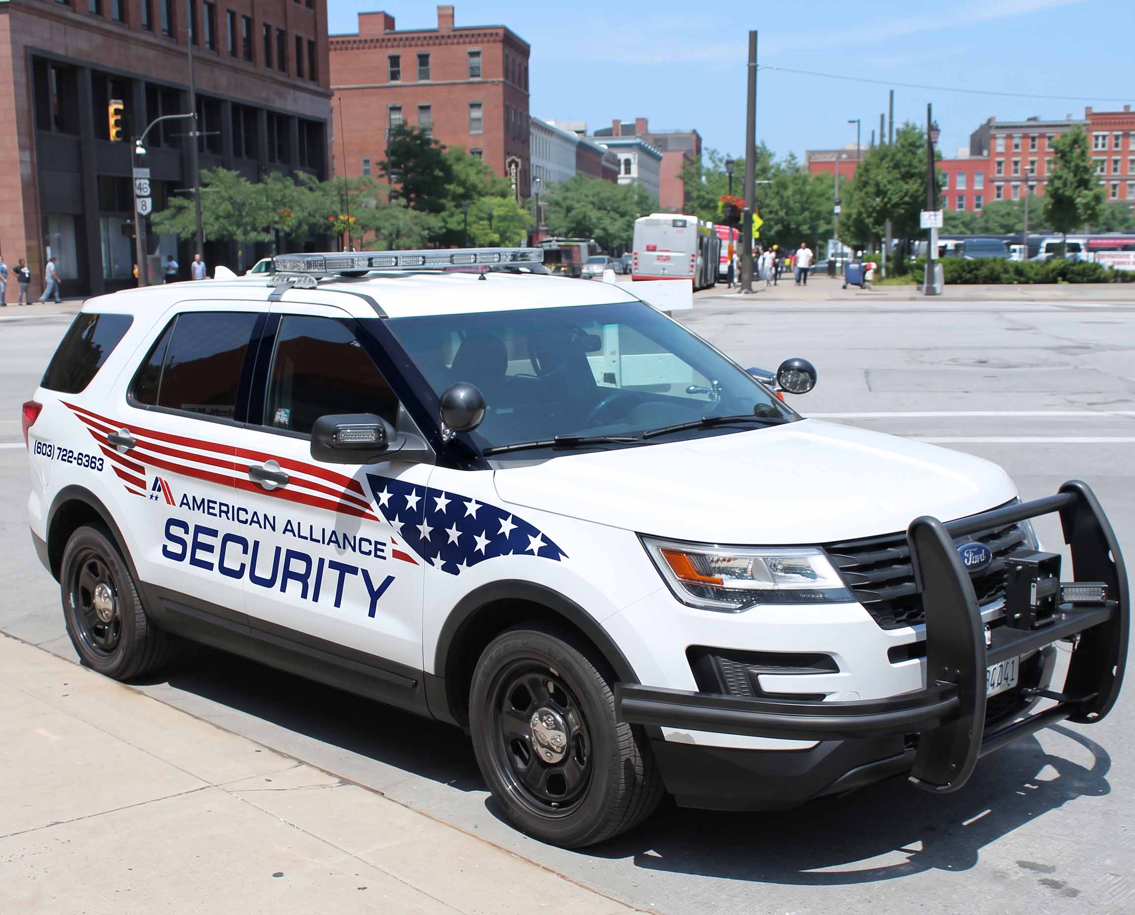 A marked American Alliance Security Ford Explorer with a roof light bar and push bumper parked on a city street